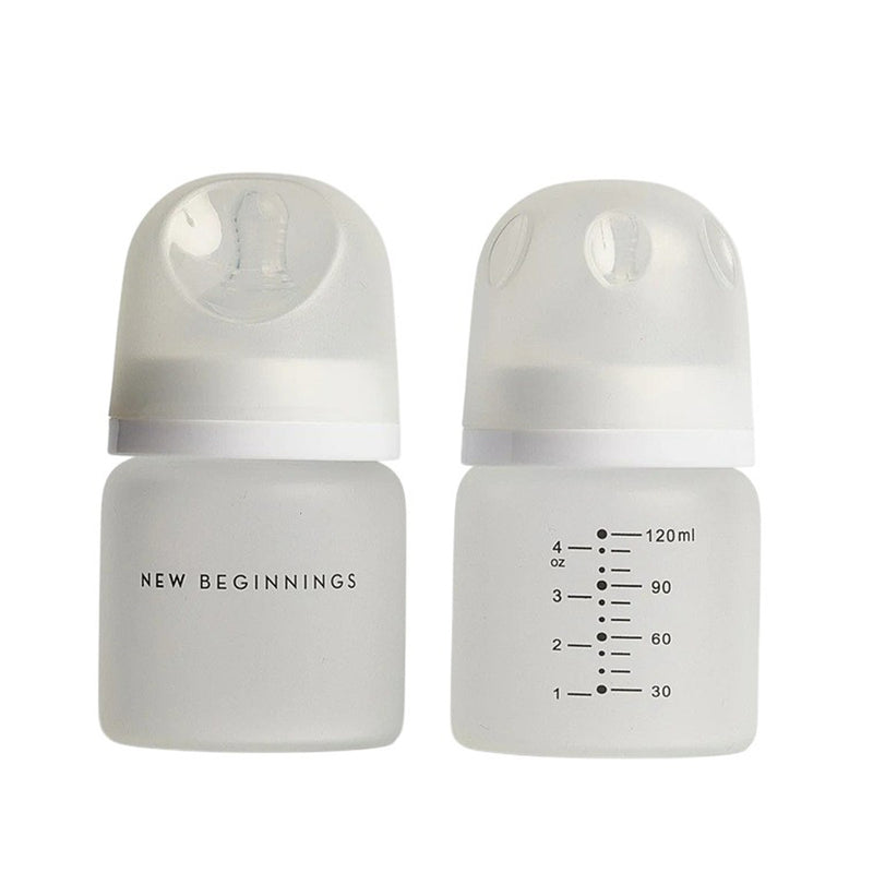 Two clear 120ml New Beginnings glass bottles with white caps and silicone nipples. One features "New Beginnings," the other has ounce and milliliter markings, both from the New Beginnings Glass Bottle 120ml Twin Silicone Outer set.