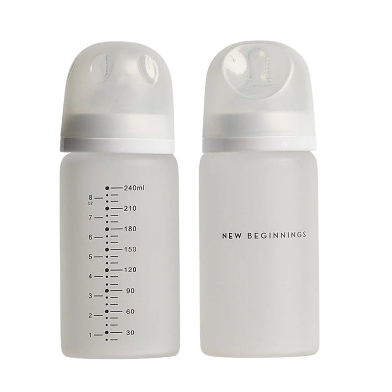 The New Beginnings Glass Bottle 240ml Twin Pack features two transparent bottles with white lids and clear caps. One has measurement markings; the other displays "NEW BEGINNINGS." Both have a protective silicone outer layer.