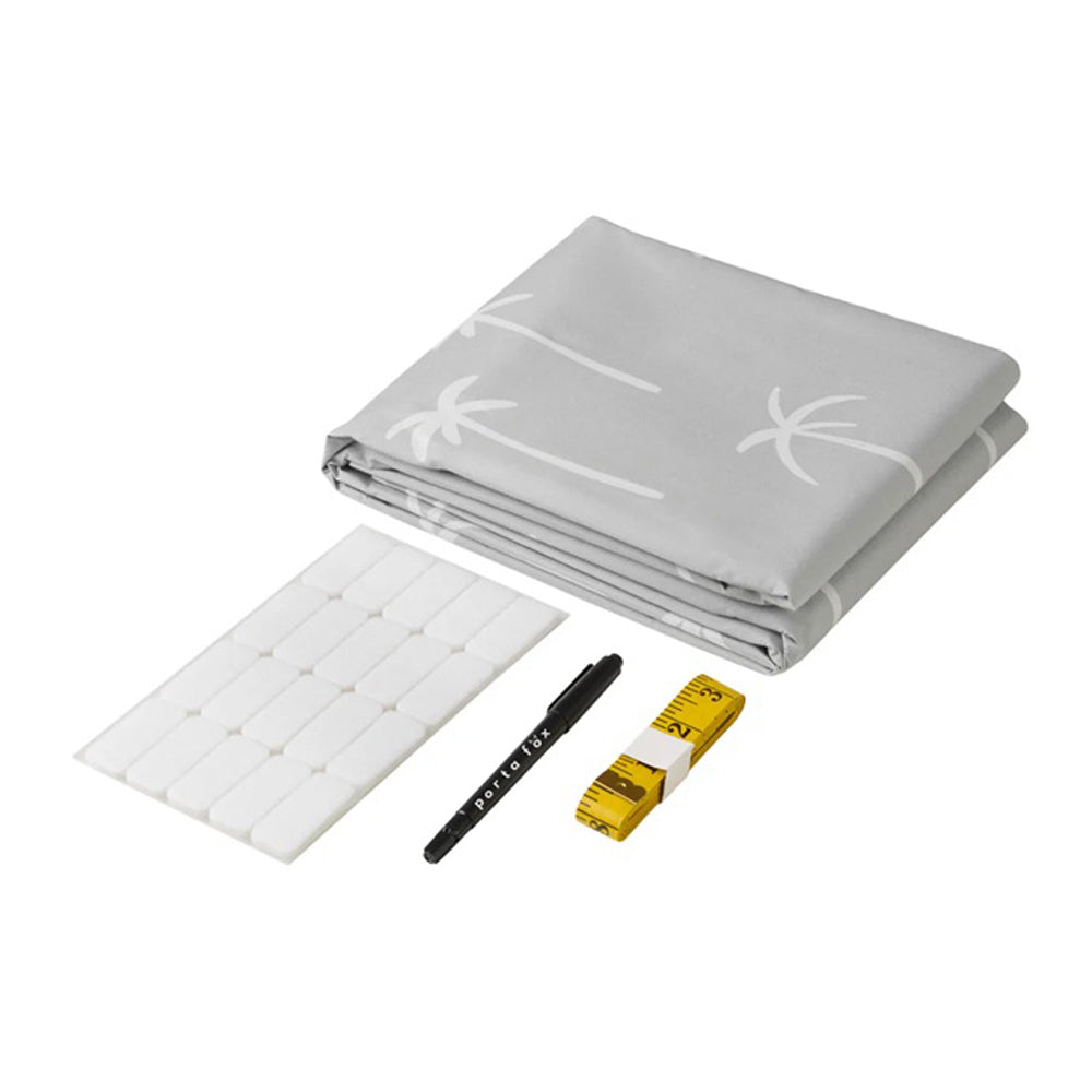 The Porta Fox Cut-to-Size Blackout Blind Boho Palms, a folded gray blind with white palm tree patterns, is shown with adhesive hooks, a black pen, and a yellow measuring tape on a white surface.