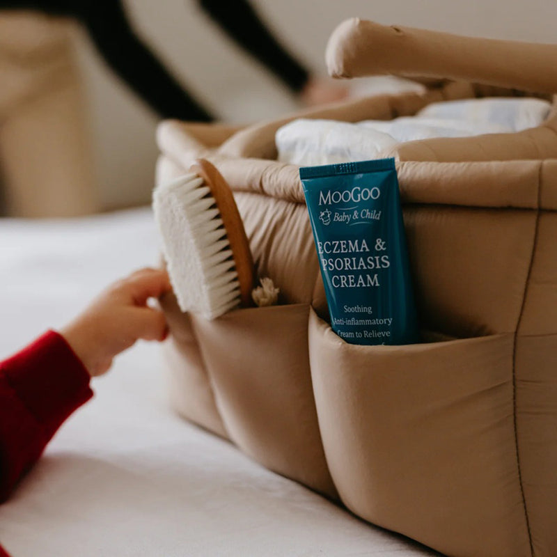 A child's hand reaches toward a Seena Migo Caddy Bag by Seena, holding a wooden brush and a blue tube of "Moogoo Eczema & Psoriasis Cream," resting on a soft, light-colored surface.