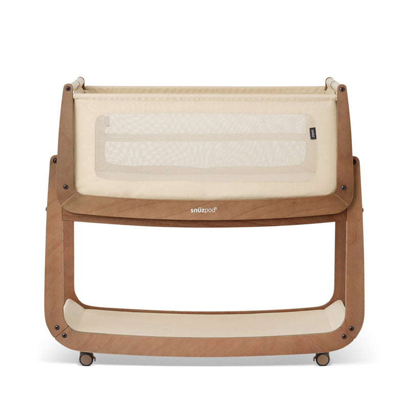 The Snuzpod 5 Bedside Bassinet Sleeper by Snuz features a wooden frame, mesh sides, fabric pocket, beige and brown colors, and small wheels for easy movement.