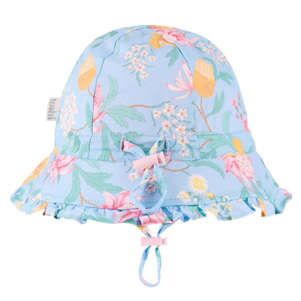 The Toshi Bell Hat Matilda Waratah Sky is a light blue cotton baby sunhat with a pink, yellow, and white floral pattern, ruffled brim, adjustable chin strap, and UPF 50+ protection—ideal for sunny days.