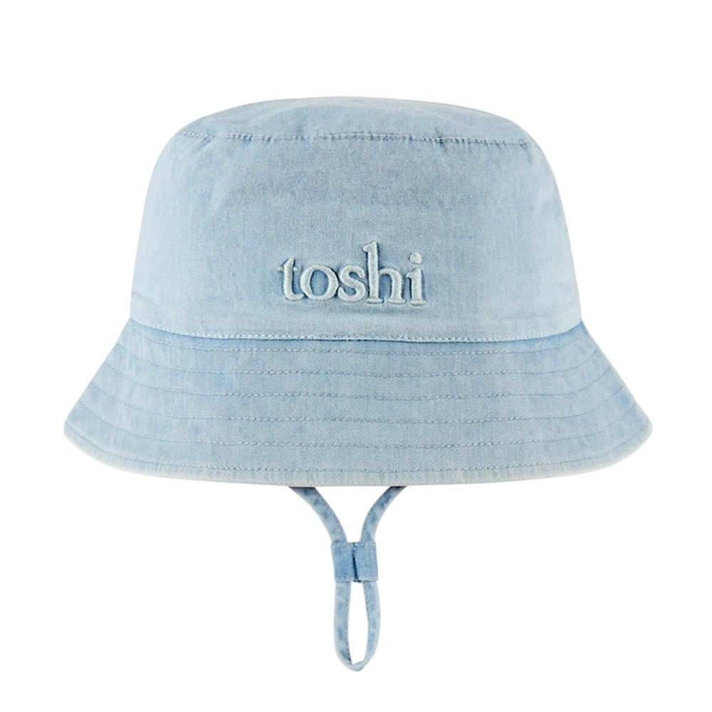 The Toshi Bucket Hat Billy Denim is a light blue denim children’s hat with "toshi" embroidered on the front, an adjustable chin strap, and built-in UPF 50+ sun protection.