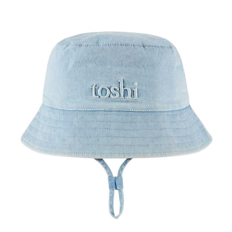 The Toshi Bucket Hat Billy Denim is a light blue denim children’s hat with "toshi" embroidered on the front, an adjustable chin strap, and built-in UPF 50+ sun protection.