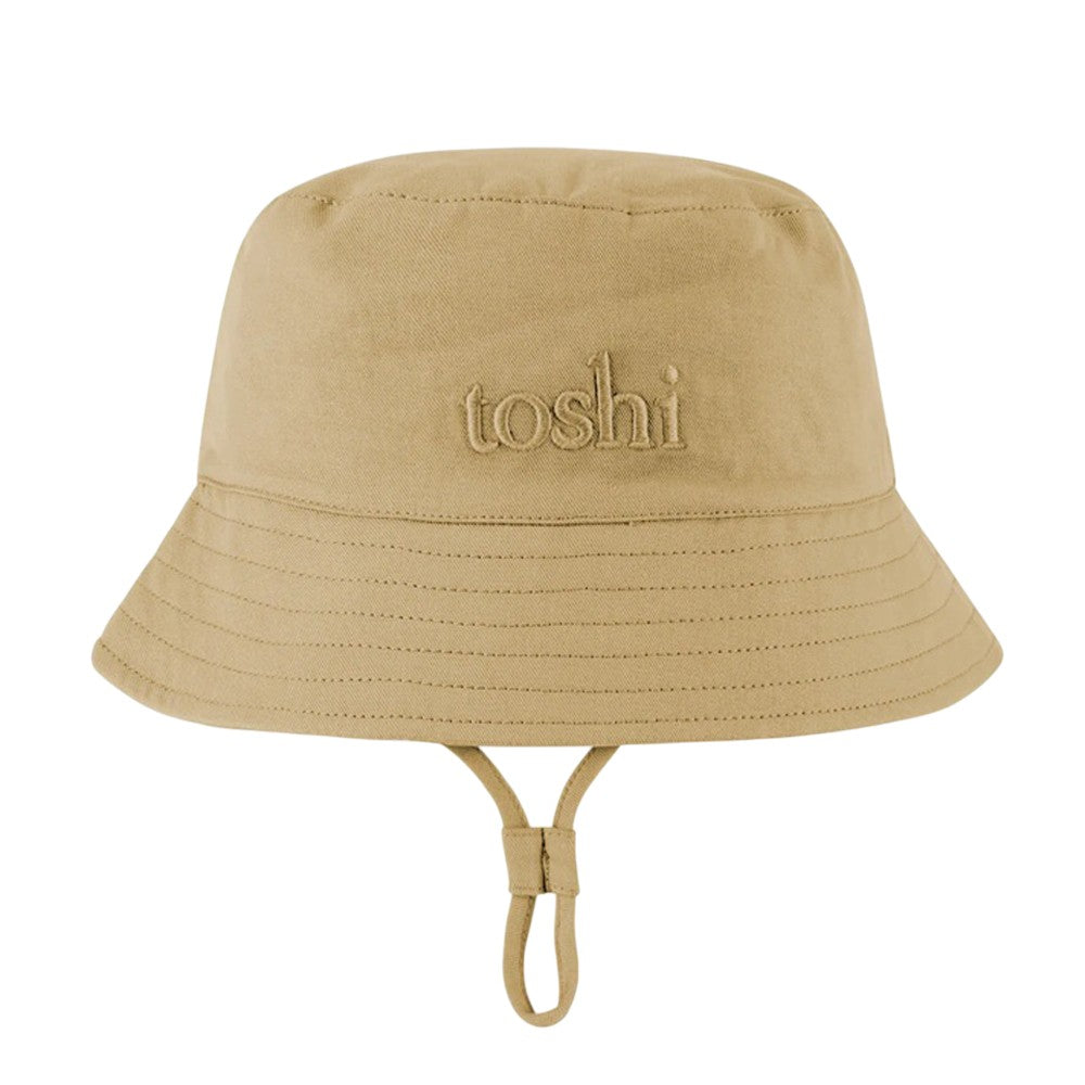 The Toshi Bucket Hat Billy Driftwood is a kids' cotton tan hat with a wide brim, visible stitching, adjustable chin strap, UPF 50+ sun protection, and "toshi" embroidered on the front.