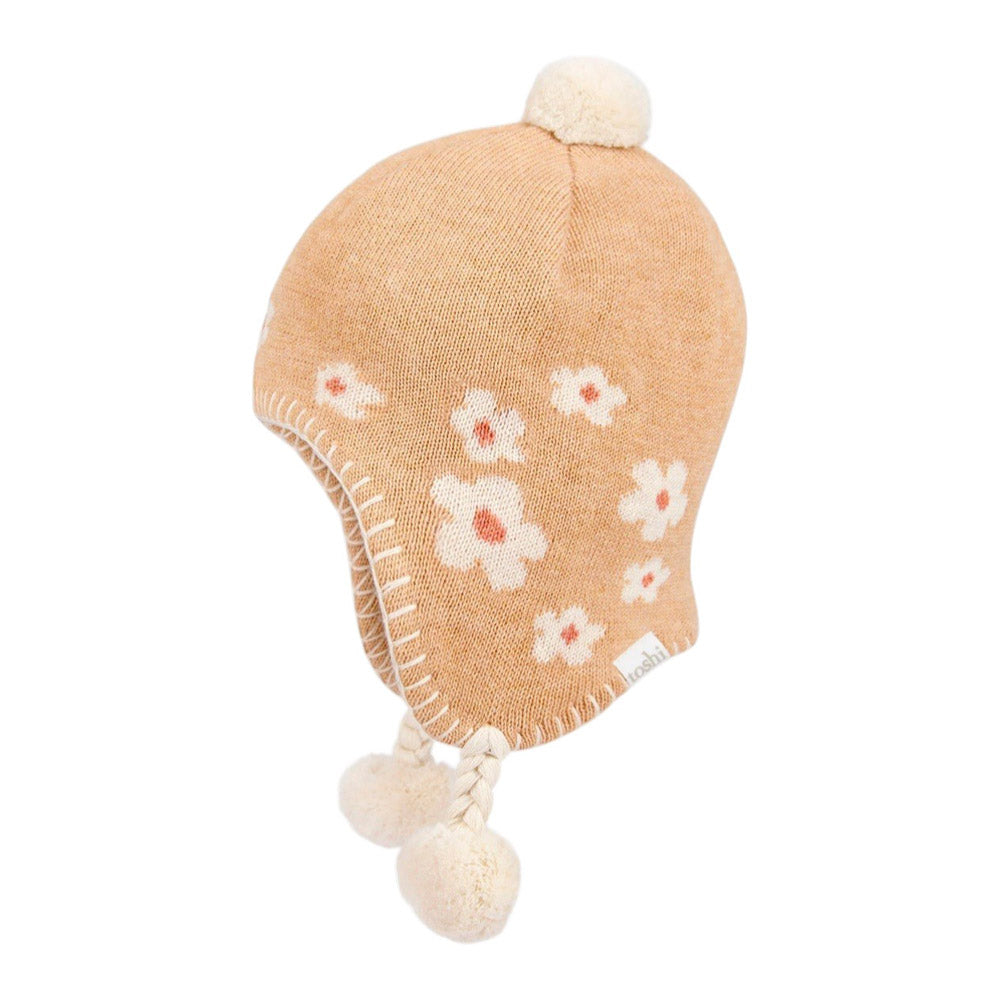 A tan knit baby winter hat featuring white and pink flower patterns, ear flaps, braided ties, and three cream pom-poms—styled like the Toshi Organic Earmuff Mystical Jasmine by Toshi.