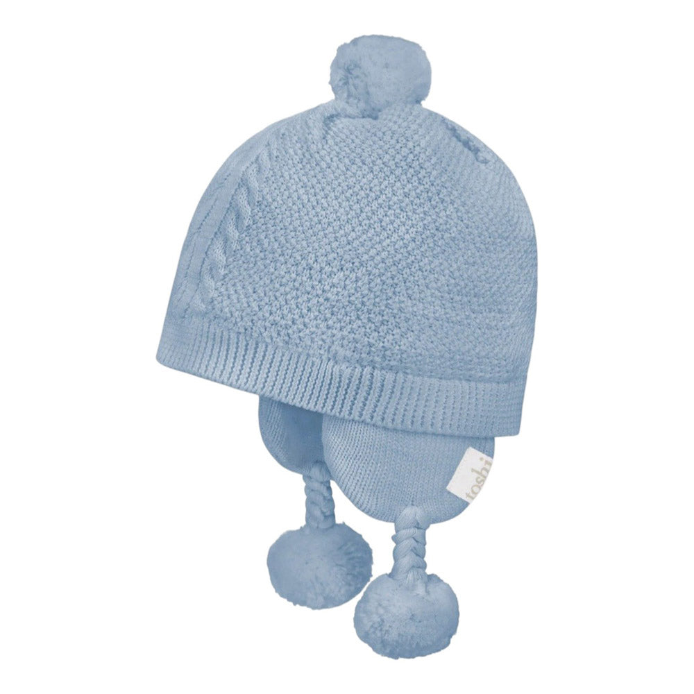 The Toshi Organic Earmuff Yummy Bluebell by Toshi is a light blue organic cotton baby hat with a pom-pom, braided ear flaps with pom-poms, textured knit pattern, and a white tag—ideal for keeping little ones cozy.