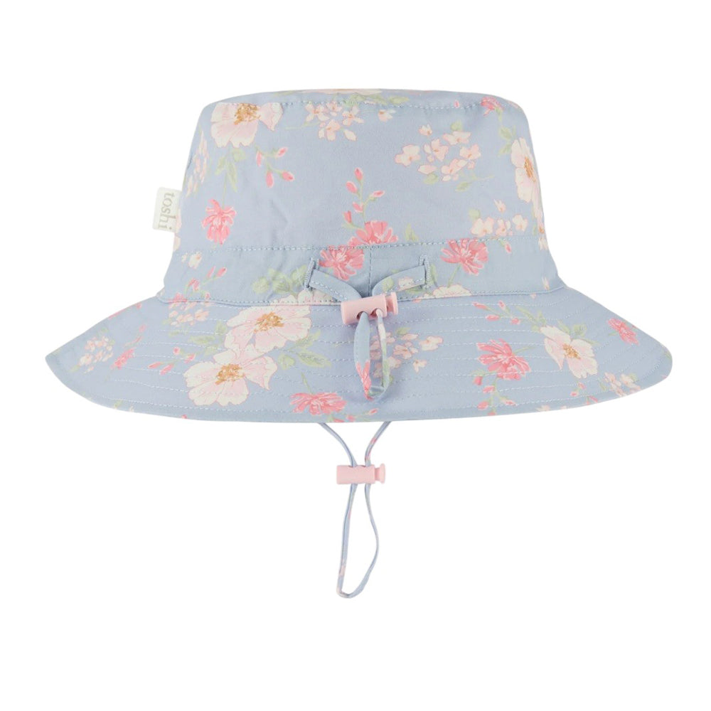 The Toshi Sunhat Desert Rose Sky by Toshi is a light blue kids’ cotton sunhat with pink and white floral patterns, a pink drawstring and bow, adjustable chin strap, and UPF 50+ for safe outdoor play.