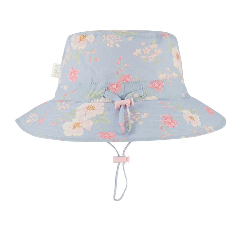 The Toshi Sunhat Desert Rose Sky by Toshi is a light blue kids’ cotton sunhat with pink and white floral patterns, a pink drawstring and bow, adjustable chin strap, and UPF 50+ for safe outdoor play.