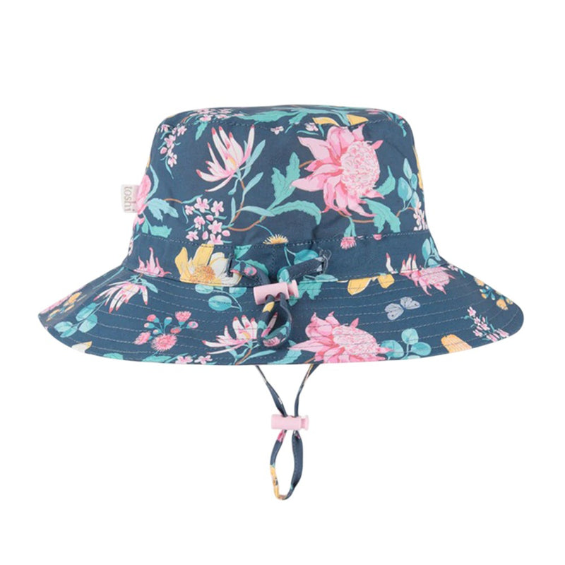 The Toshi Sunhat Waratah Midnight is a navy kids hat with pink and yellow floral print, wide brim, adjustable chin strap, pink toggle, and UPF 50+—ideal sun protection for children.