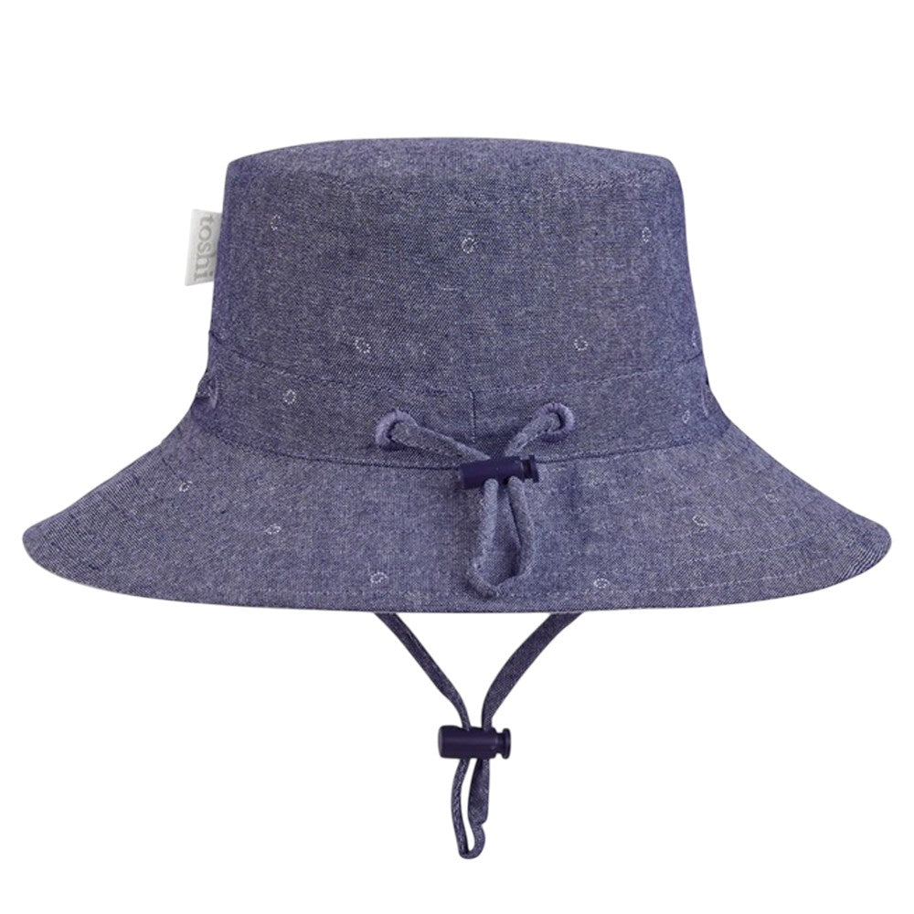 The Toshi Sunhat XavierInk is a purple children’s bucket hat with UPF 50+ sun protection, wide brim, adjustable chin strap, subtle patterned fabric, a front bow, and side tag.