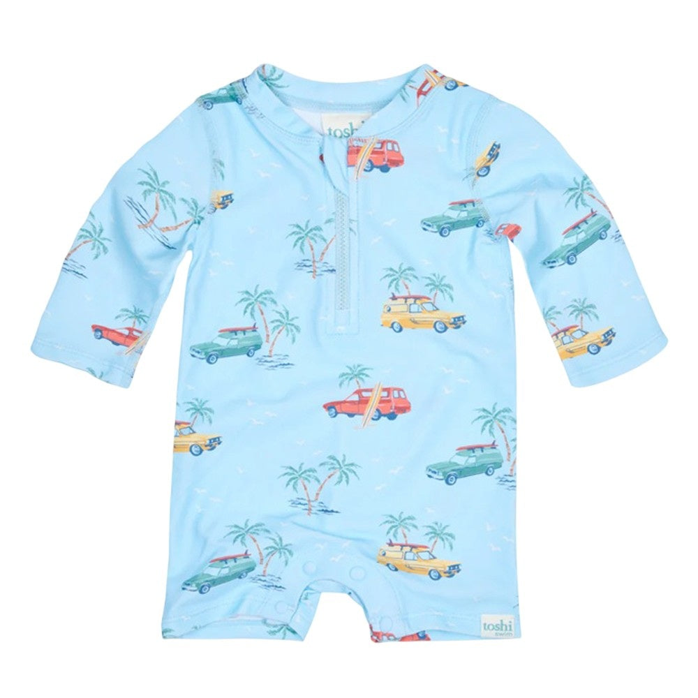 The Toshi Swim Onesie L/S Splash Swell Seekers Sky by Toshi is a light blue baby romper with long sleeves, UPF 50+ sun protection, and playful prints of vintage cars, palm trees, and surfboards. Features snaps at the legs and buttons at the collar.