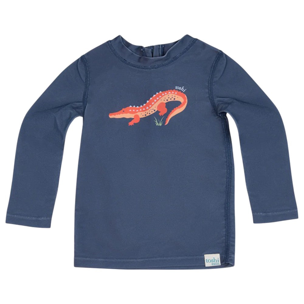 The Toshi Swim Rashie L/S Splash Kakadu Moonlight is a navy long-sleeve children’s shirt featuring an orange alligator graphic and UPF50+ sun protection, ideal as baby swimwear from the trusted brand Toshi.