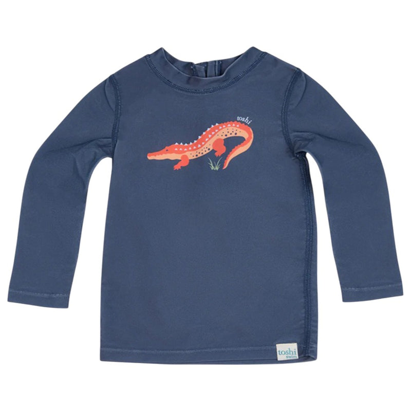 The Toshi Swim Rashie L/S Splash Kakadu Moonlight is a navy long-sleeve children’s shirt featuring an orange alligator graphic and UPF50+ sun protection, ideal as baby swimwear from the trusted brand Toshi.