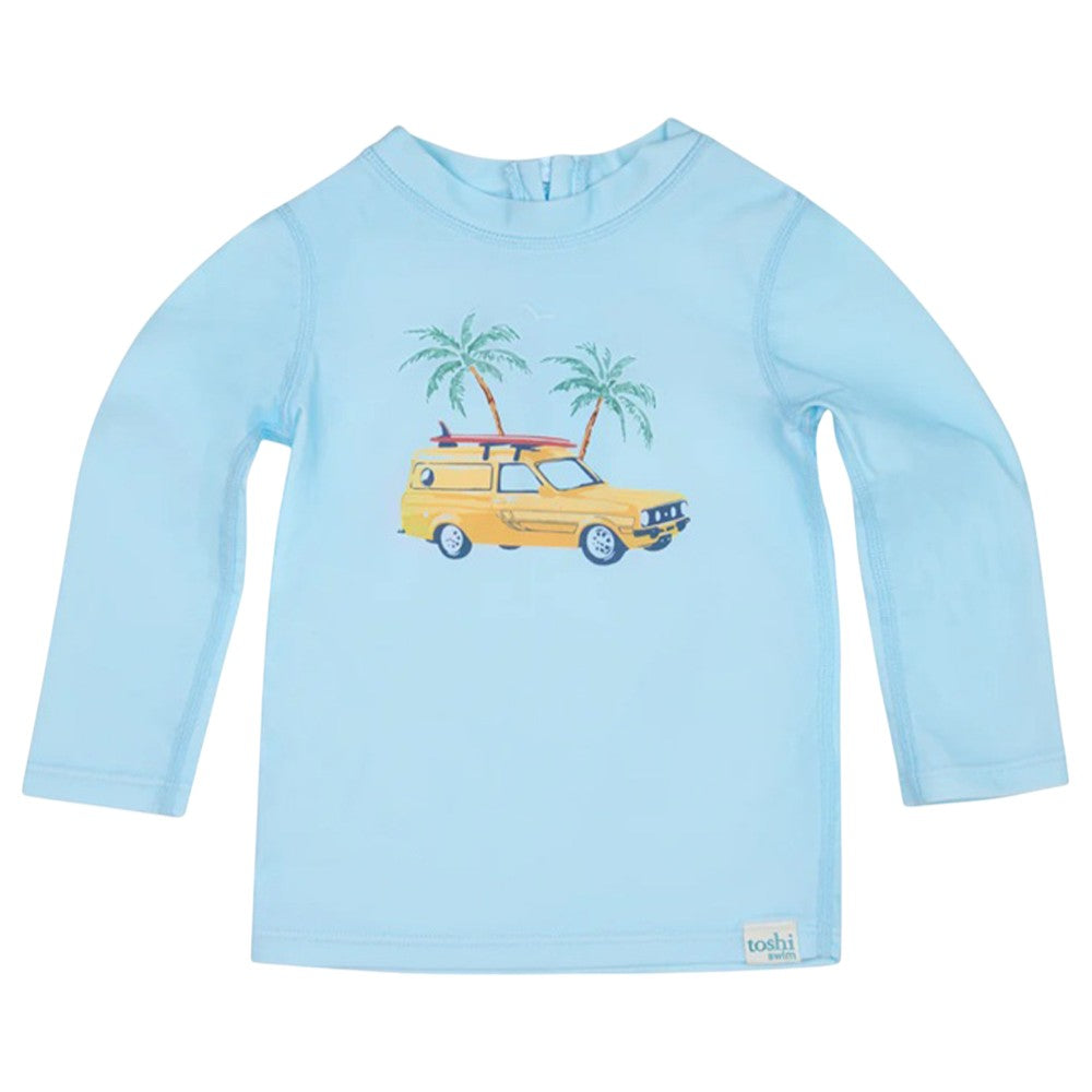 The Toshi Swim Rashie L/S Splash Swell Seekers Sky is a light blue long-sleeve baby rash guard with a yellow station wagon, surfboard, and palm tree print. Featuring UPF50+ protection and a "toshi" label at the hem—ideal for sunny days.