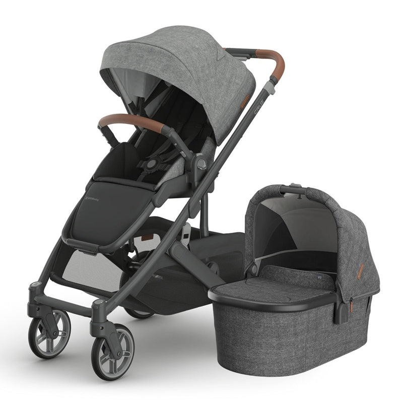 The UPPAbaby Cruz V3 stroller in modern gray with a brown handlebar and matching UPPAbaby V3 Bassinet, both featuring a sleek design and black wheels, are shown side by side.