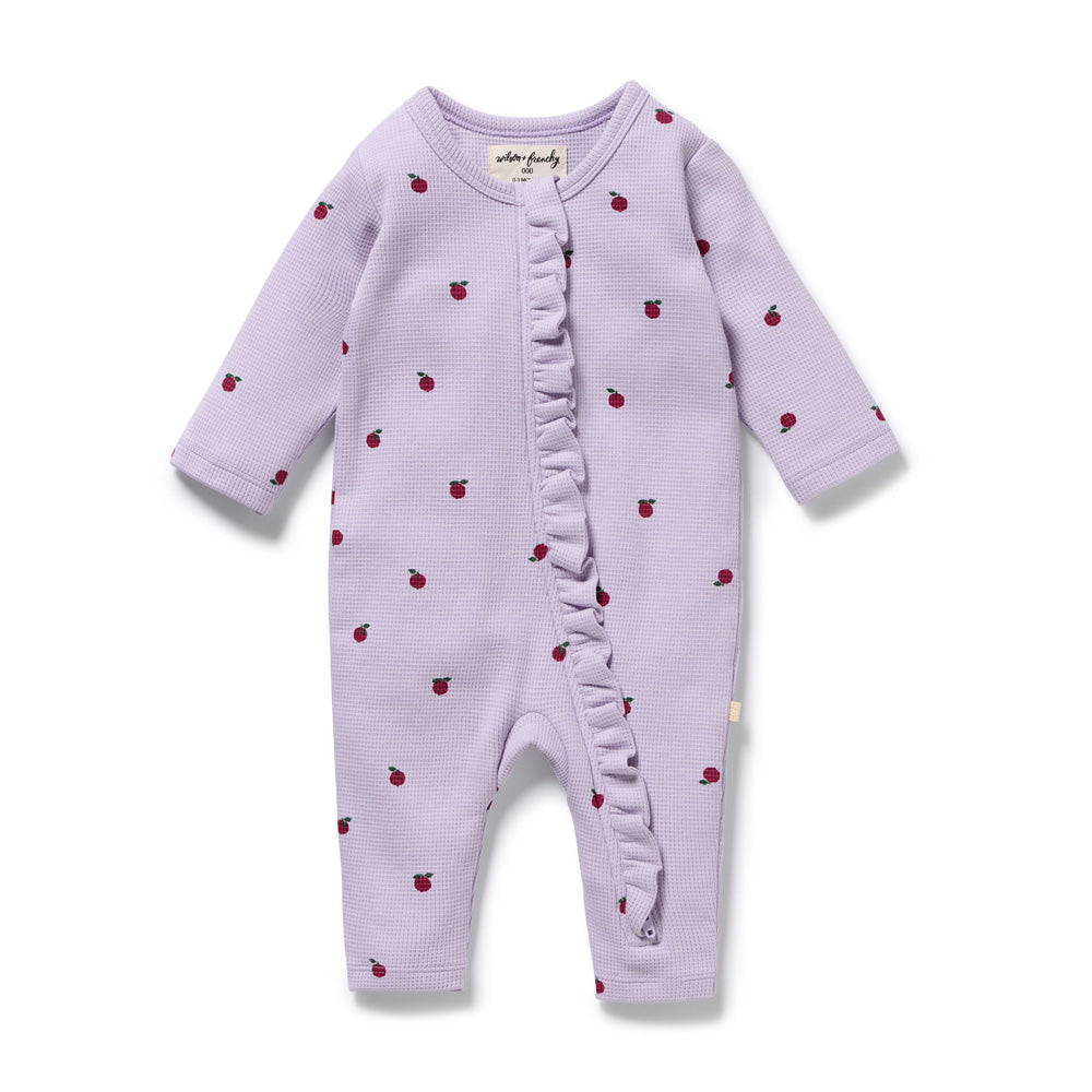 The Wilson & Frenchy Berry Plums Organic Waffle Ruffle Zipsuit is a lavender long-sleeve baby romper with small red plum prints, diagonal ruffle detail, round neckline, and snaps along the inseam.