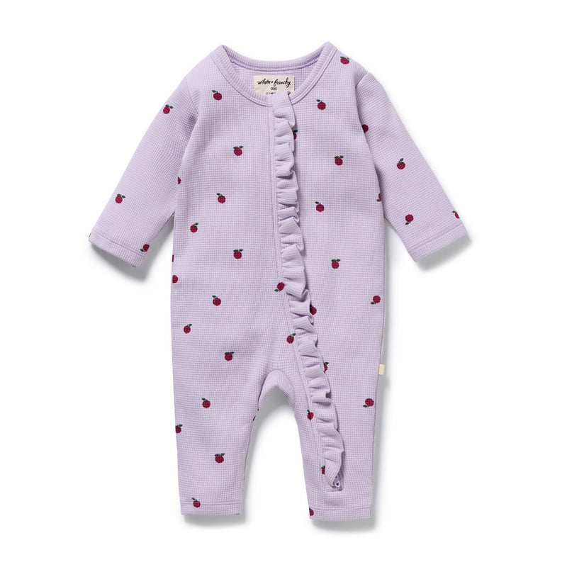 The Wilson & Frenchy Berry Plums Organic Waffle Ruffle Zipsuit is a lavender long-sleeve baby romper with small red plum prints, diagonal ruffle detail, round neckline, and snaps along the inseam.