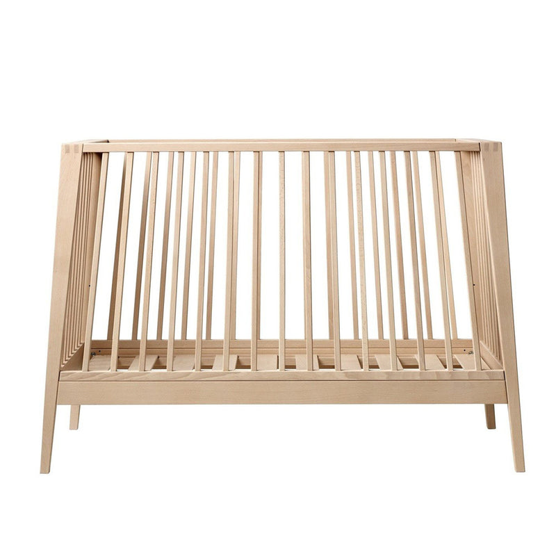 The Leander Linea Cot by Leander features angled legs, vertical slats on all sides, and a minimalist modern design. Shown empty against a plain white background, this natural wood crib easily converts into a toddler bed.
