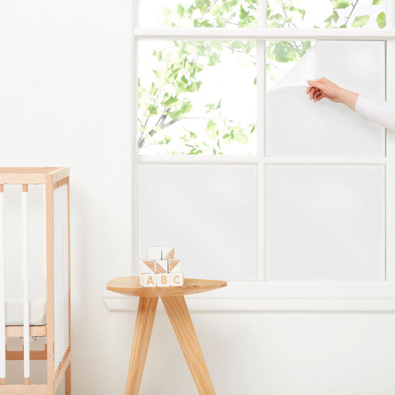 A hand peels ErgoPouch Window Blockout, a reusable static-cling frosted film from ErgoPouch, off a bright window beside a wooden crib and table with ABC blocks. Green leaves are visible outside the blackout covering.