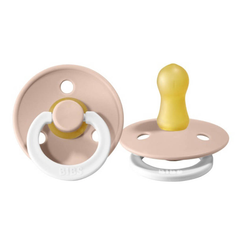 Two beige and white Bibs Dummies BIBS Colour Soothers Night Glow pacifiers are shown, each with a round shield, ring handle, and yellow natural rubber nipple. One faces front; the other is angled to highlight the nipple.