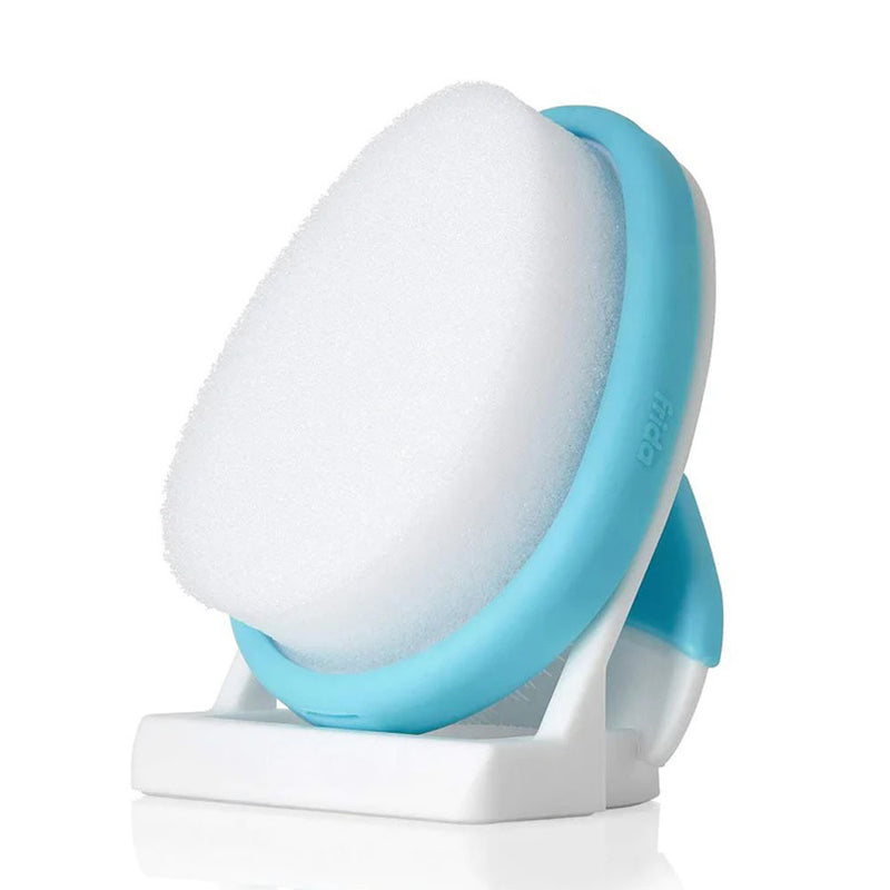 A close-up of the Frida Baby Flake Fixer, a white oval sponge with a light blue handle by Frida Baby, designed for gentle baby hair care and cradle cap relief, shown resting on its matching stand against a white background.