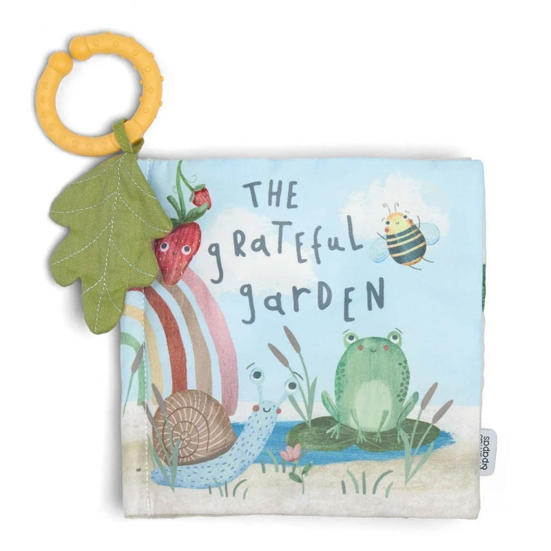 The Mamas & Papas Grateful Garden Interactive Activity Book is a soft fabric baby book featuring frog, snail, bee, and strawberry characters, plus a yellow textured teething ring and green leaf attached.