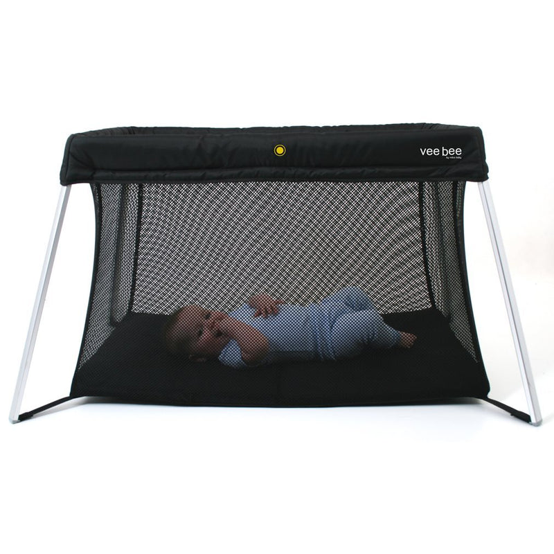 A baby in a striped onesie lies on their side inside a black mesh Vee Bee Amado Travel Cot with silver legs, featuring the "vee bee" label in the top right corner.