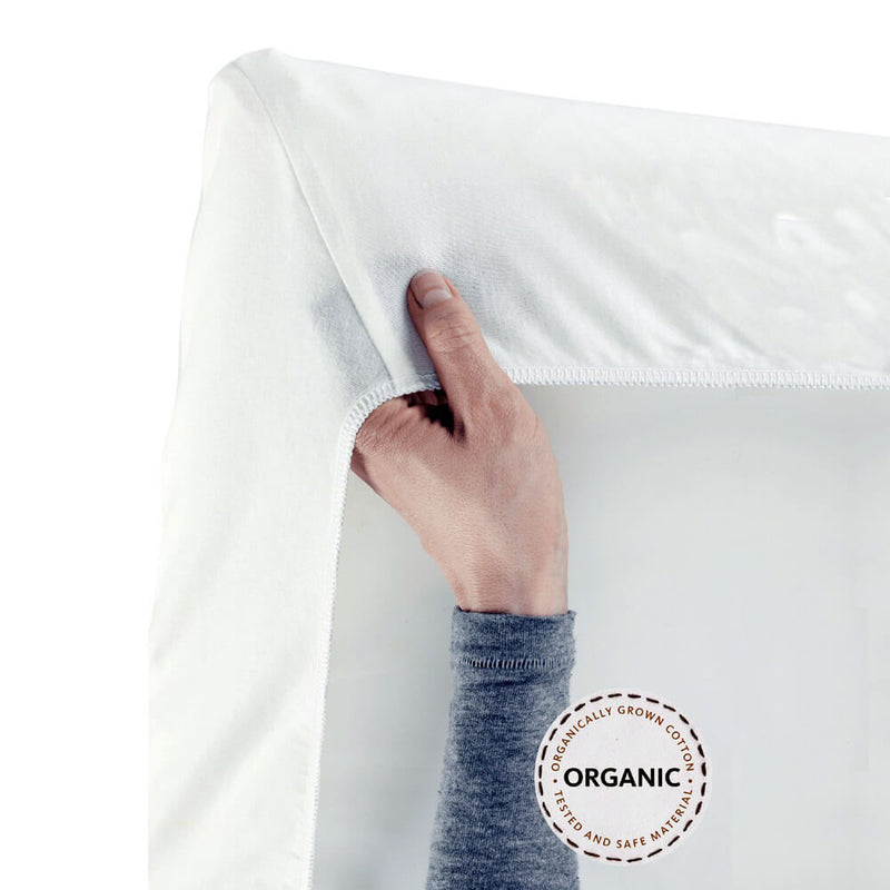 A hand lifts the corner of a white BabyBjorn Travel Cot Fitted Sheet, showing its snug stretchy fit on a BabyBjorn crib mattress. A circular badge reads "ORGANIC - CERTIFIED AND SAFE MATERIALS.